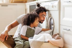 Cheerful black kid boy sitting on dad's lap and helping father at linen in basket while doing laundry near washing machine in flight kitchen in weekend at home